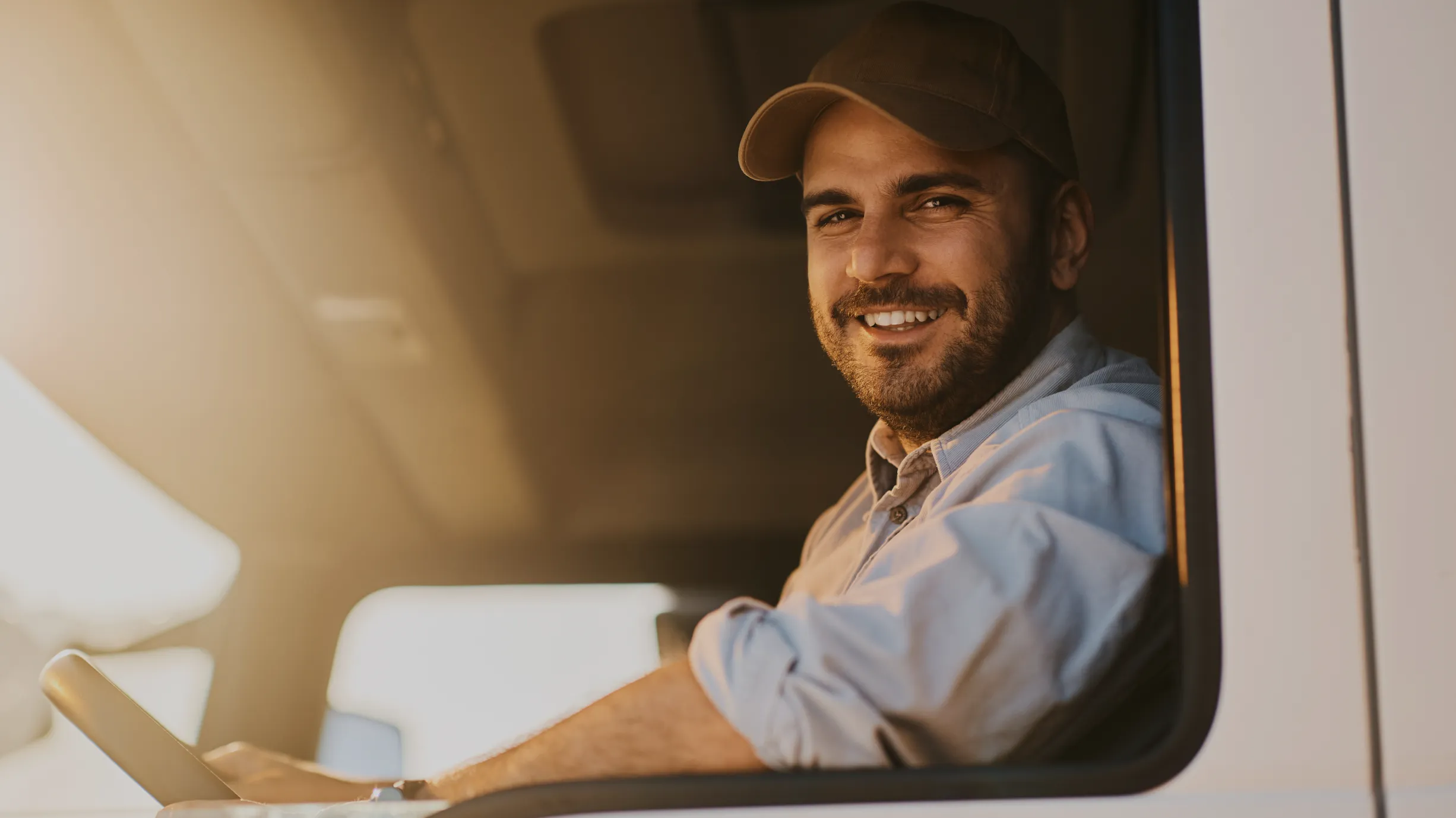 A smiling delivery driver in a cap at the wheel of his van, arm resting on the open window